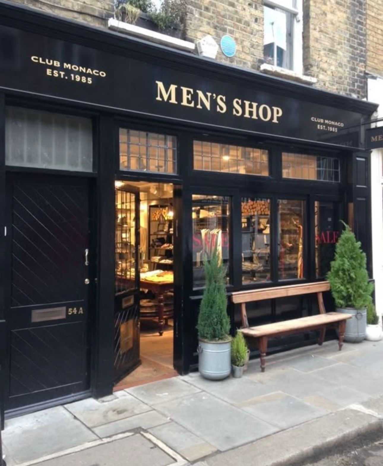 Exterior of a vintage-style men's clothing shop with a wooden bench and potted plants outside.