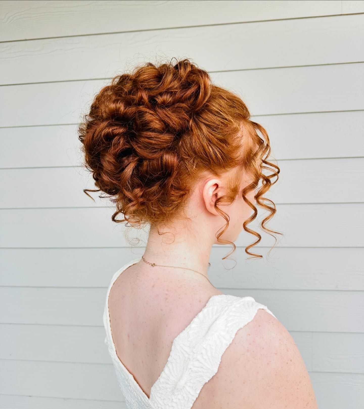 Woman with curly red hair styled in an elegant updo, wearing a white dress against a light background.